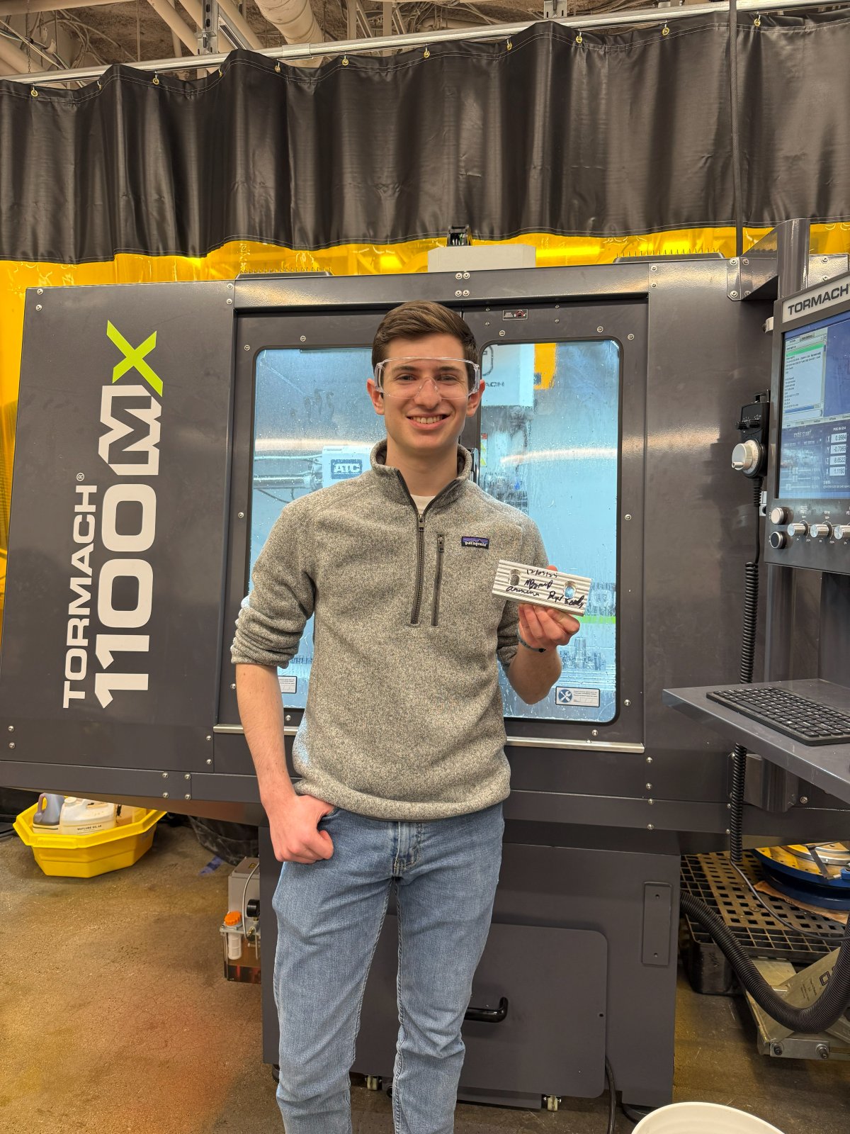 Noah in safety glasses holding a machined aluminum part in front of a Tormach 1100MX CNC mill.