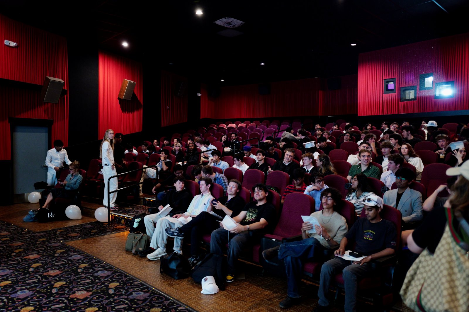 A packed cinema with red walls and dark red seats, students filling most of the rows.