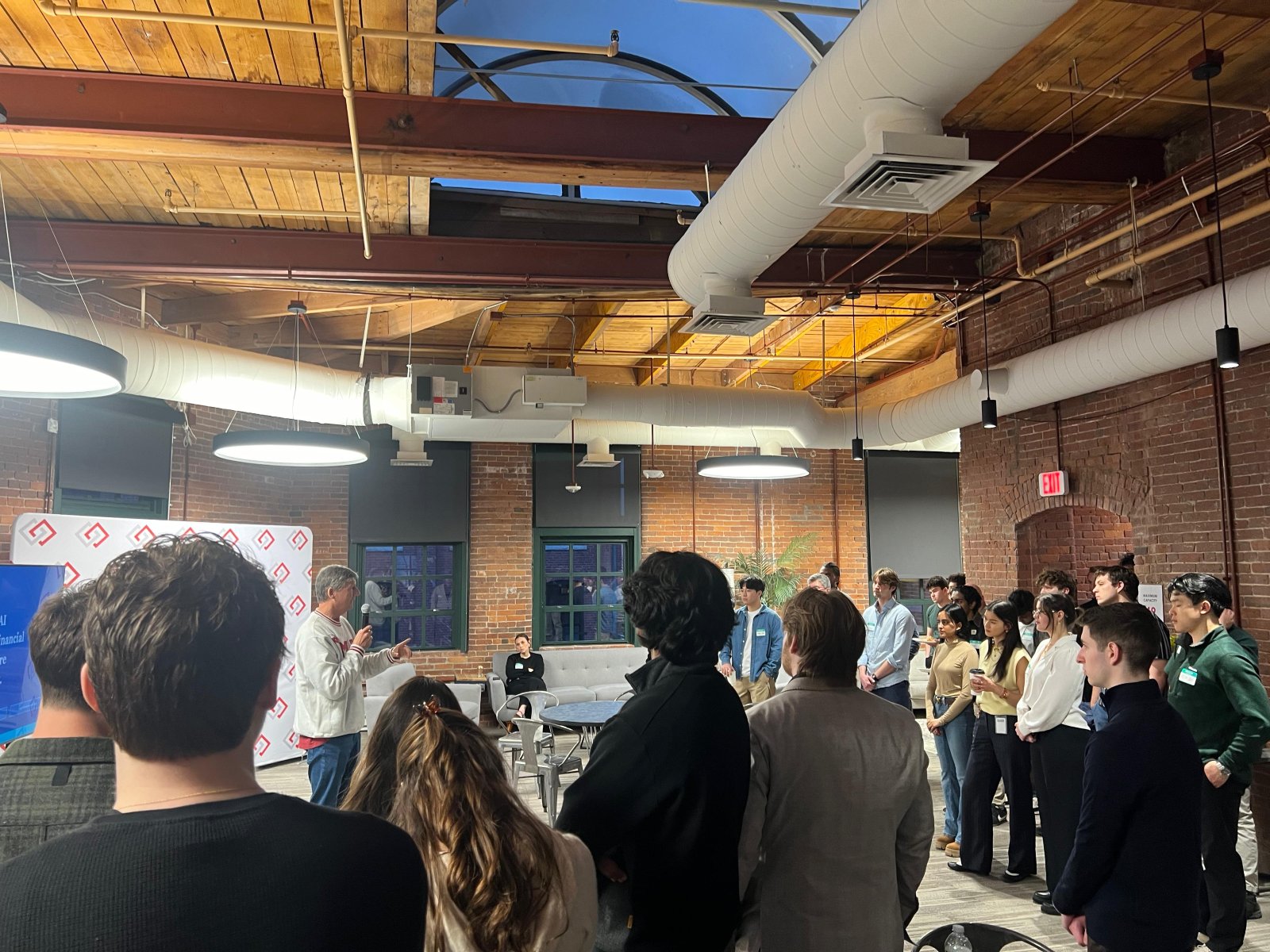 A crowd standing in a brick-and-timber loft watching a speaker at a pitch event.