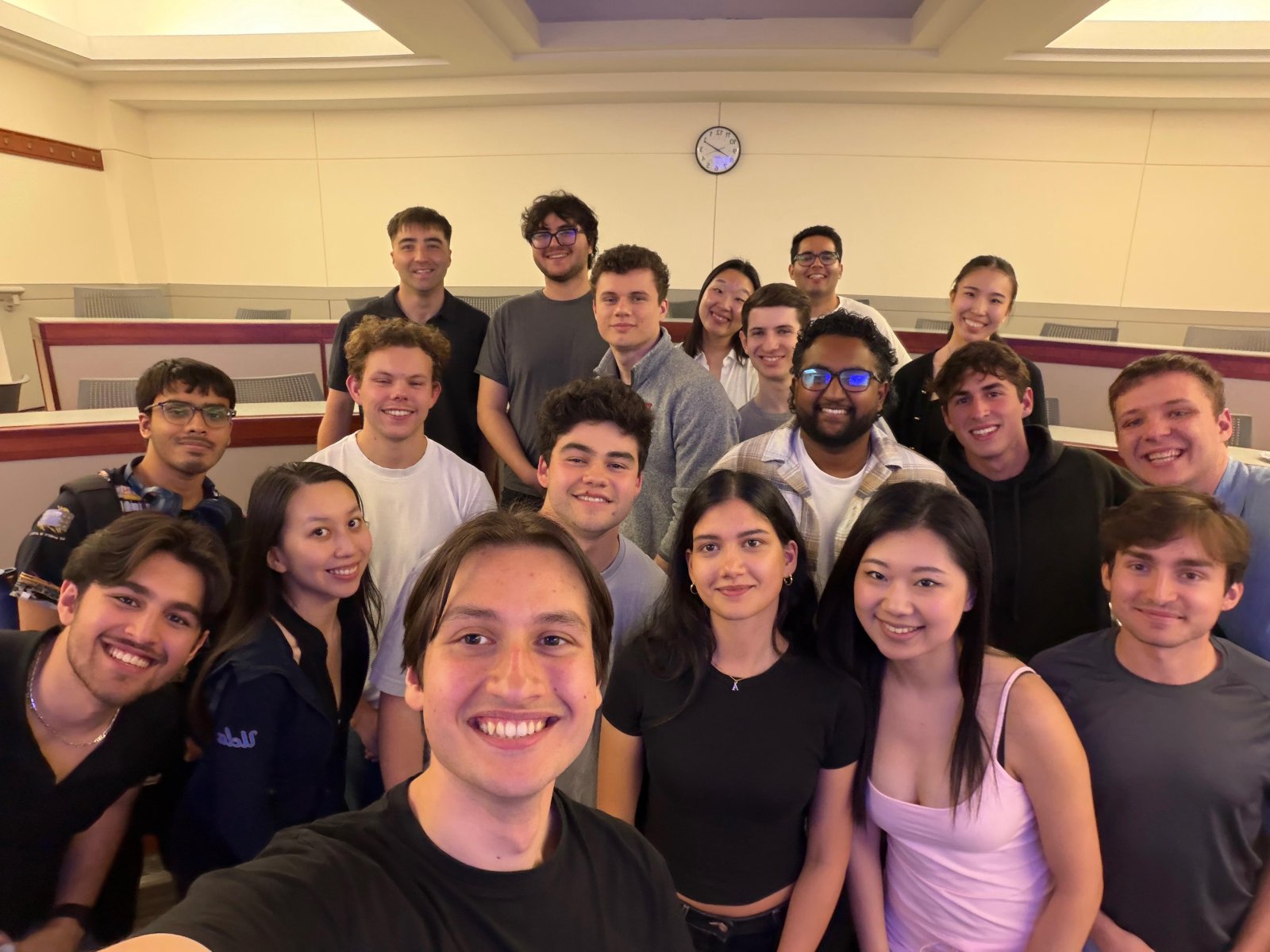 A group of about sixteen people crowded together for a photo in a tiered lecture room.