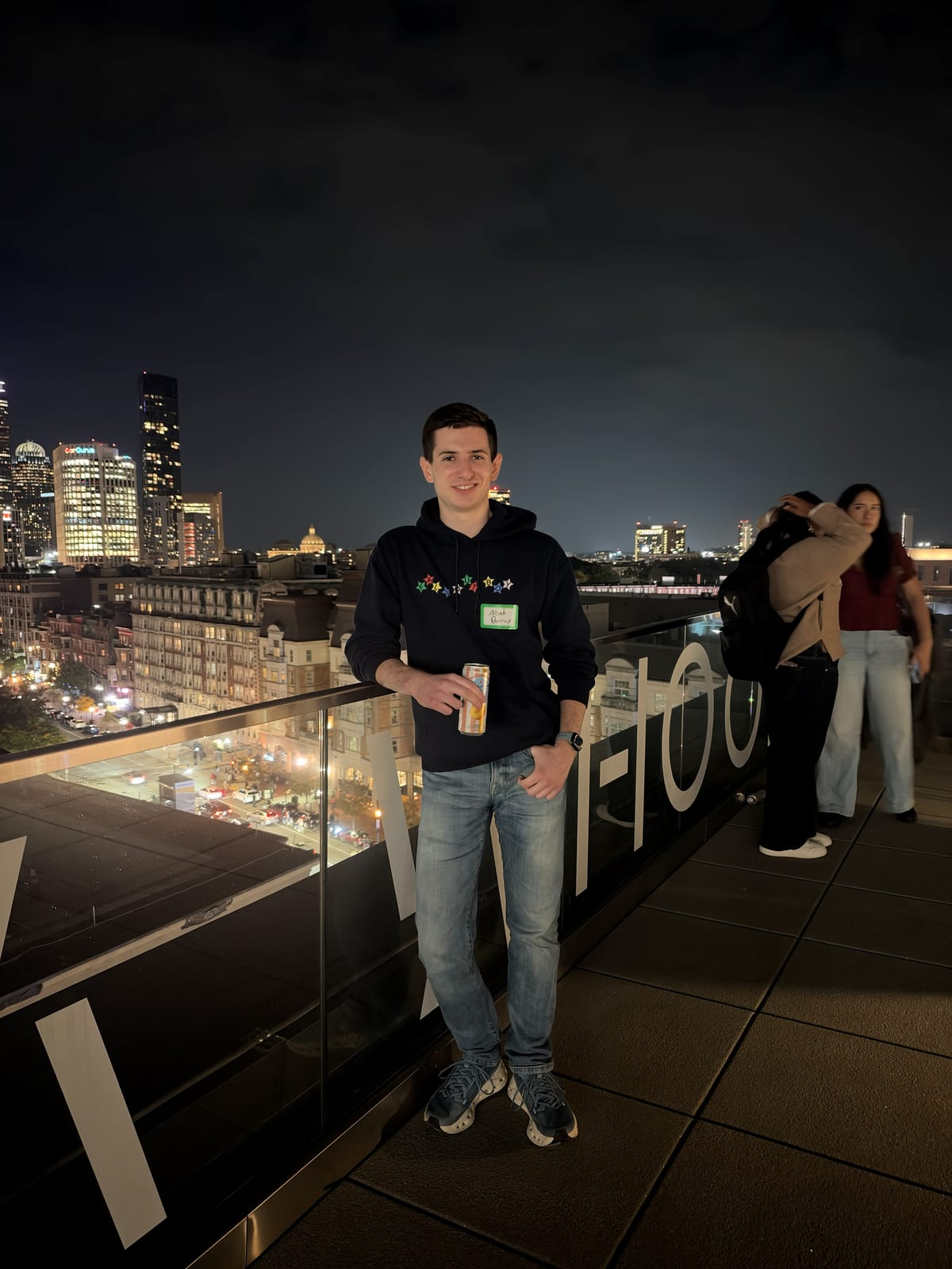 Noah on a rooftop terrace at night, leaning on a glass railing with the Boston skyline lit up behind him.